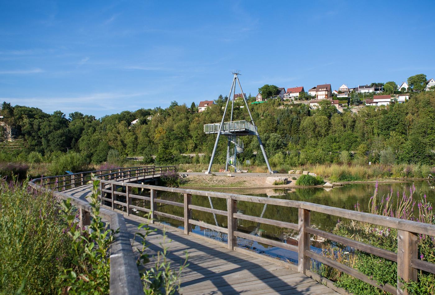 Holzsteg über ein Gewässer mit einem Aussichtsturm im Hintergrund.