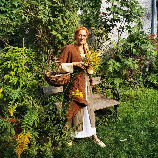 Eine rothaarige Frau in einem braunen Umhang gekleidet, steht in einem Garten. Über dem Arm trägt sie einen Korb und in der Hand hält sie gelb blühende Pflanzen.