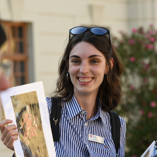 Eine Frau mit Sonnebrille auf dem Kopf und braunen Haaren lacht in die Kamera und hält ein Bild hoch.
