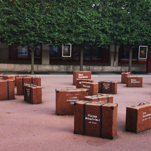 Auf einem rötlichen Untergrund, wo einst die Synagoge stand, stehen Koffer mit Namen darauf.