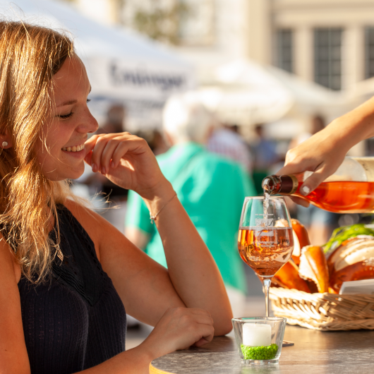 Zu sehen ist eine lächelnde Frau, der ein Glas Wein eingeschenkt wird.